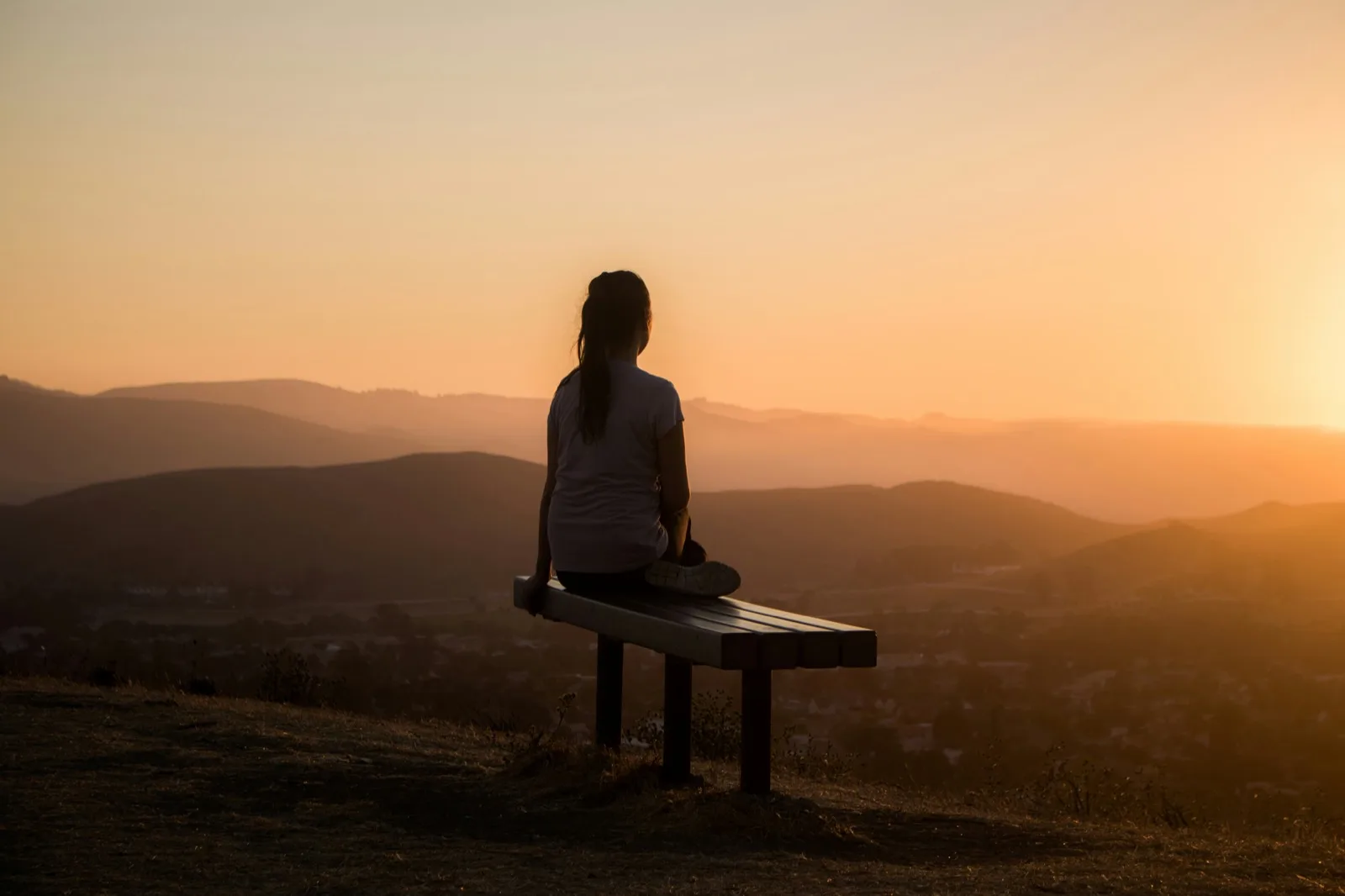 A woman sitting alone on a hilltop bench, looking out over layered hills at sunset